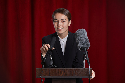 Portrait of young woman speaking to microphone on stage standing at podium against red curtain, copy space