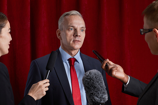 Waist up portrait of mature man giving interview to journalist and speaking to microphones against red curtain