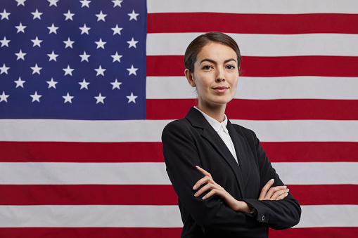 Waist up portrait of confident female politician looking at camera while standing with arms crossed against USA flag background, copy space