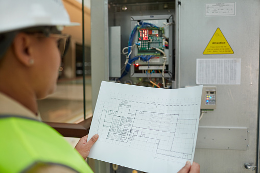 Female worker holding blueprints while inspecting electrical switchboard at construction site, copy space