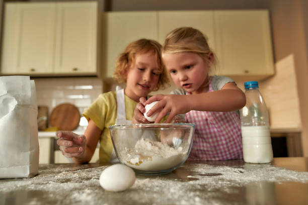 istock Two cute little children, boy and girl in aprons looking focused while adding eggs, preparing dough together on the kitchen table at home 1293449076