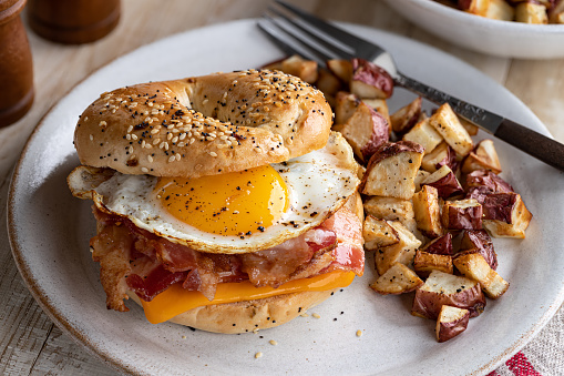 Bagel breakfast sandwich with fried egg, bacon and cheese on a plate with potatoes
