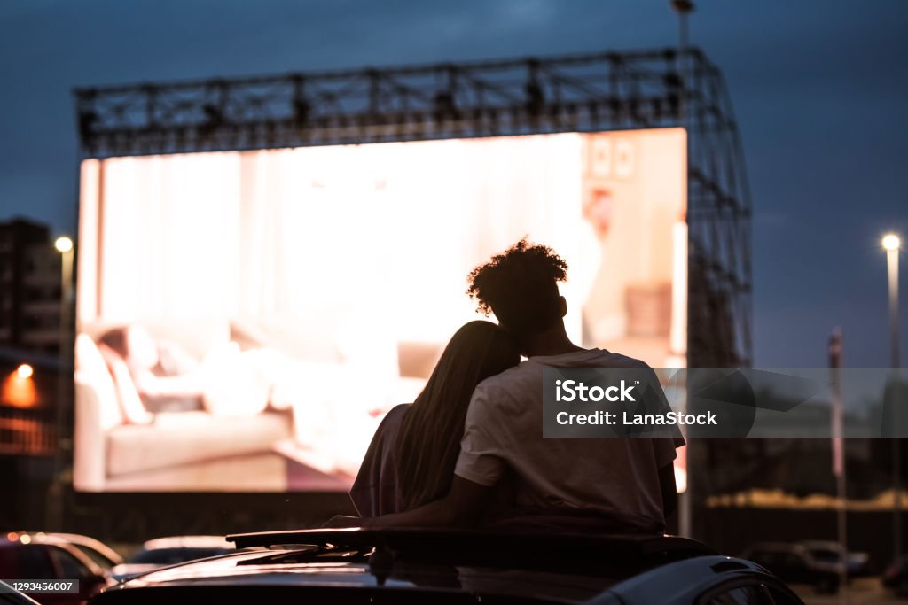 Silhouetted view of attractive young couple, boy and girl embracing, spending time together, sitting in the car while watching a movie in a drive in cinema Silhouetted view of attractive young couple, boy and girl embracing, spending time together, sitting in the car while watching a movie in a drive in cinema. Entertainment, dating concept. Rear view Movie Stock Photo