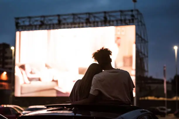 Silhouetted view of attractive young couple, boy and girl embracing, spending time together, sitting in the car while watching a movie in a drive in cinema. Entertainment, dating concept. Rear view