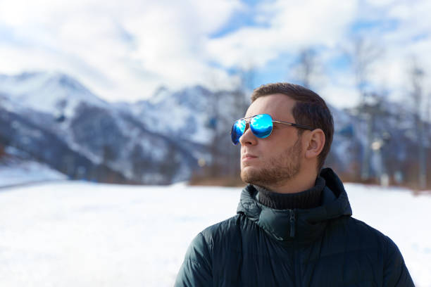 Stylish young man in down jacket and sunglasses standing in front of ski lifts and snow mountains panorama covered in snow. Confidently looking away on territory of winter resort near Sochi, Russia Stylish young man in down jacket and sunglasses standing in front of ski lifts and snow mountains panorama covered in snow. Confidently looking away on territory of winter resort . High quality photo sochi photos stock pictures, royalty-free photos & images