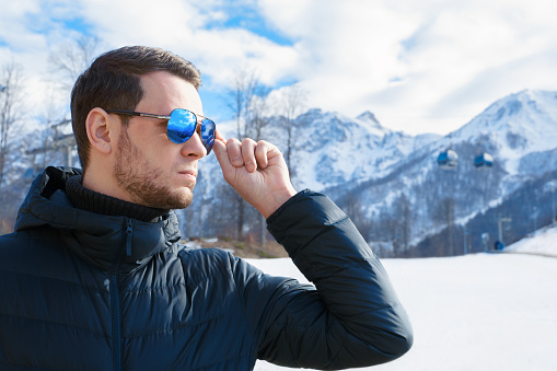 Stylish young man in down jacket and sunglasses standing in front of ski lifts and snow mountains panorama covered in snow. Confidently looking away on territory of winter resort . High quality