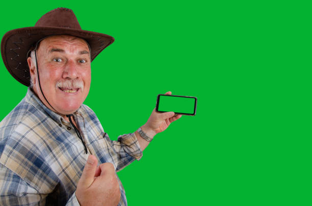 The elderly man in a plaid shirt and brown hat rejoices at the good news received by the phone. Shot on a green background.