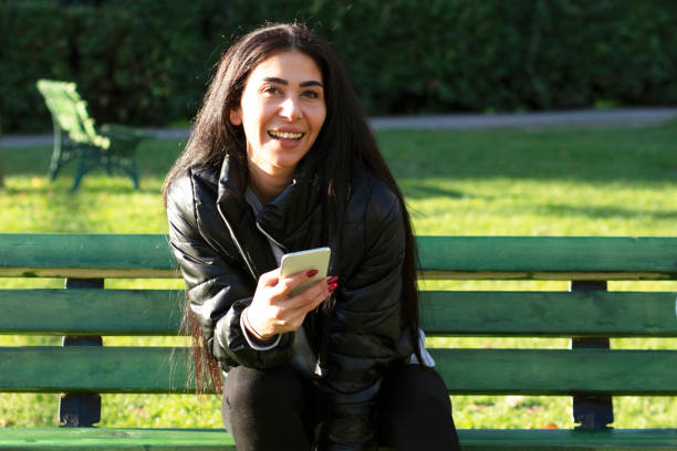istock Young woman sitting on a bench in a public park using mobile phone 1293475933