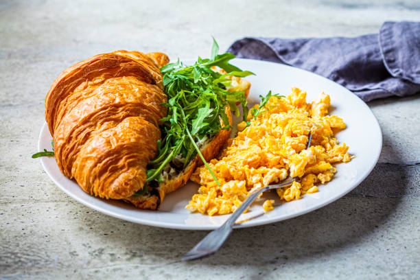 istock Scrambled eggs and croissant with cheese and arugula on white plate. Eggs and croissant for breakfast. Breakfast concept. 1293479472