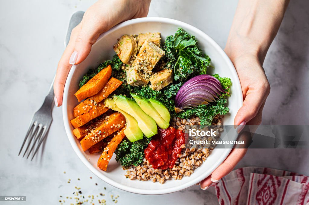 Woman hands eating vegan salad of baked vegetables, avocado, tofu and buckwheat buddha bowl, top view. Plant based food concept. Baked vegetables, avocado, tofu and buckwheat buddha bowl. Vegan lunch salad with kale, baked sweet potato, tofu, buckwheat and avocado in a white bowl. Vegan concept. Healthy Eating Stock Photo