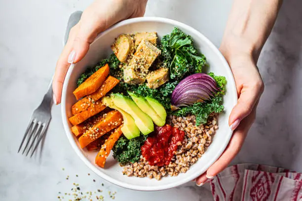 Photo of Woman hands eating vegan salad of baked vegetables, avocado, tofu and buckwheat buddha bowl, top view. Plant based food concept.