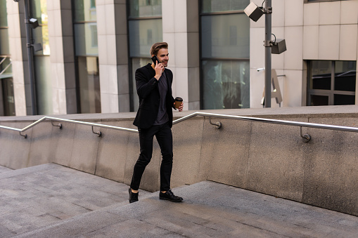 Confident young manholding coffee cup and talking on mobile phone while walking outdoors