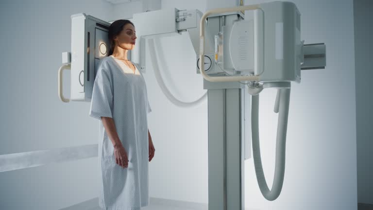 Hospital Radiology Room: Beautiful Multiethnic Woman in Medical Gown Standing Next to X-Ray Machine while Female Doctor Adjusts it. Healthy Patient Undergoes Routine Scanning with the Nurse's Help.