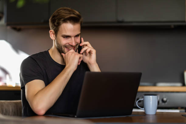 istock Young handsome man working from home in the kitchen with a laptop talking on mobile and smile 1293482512