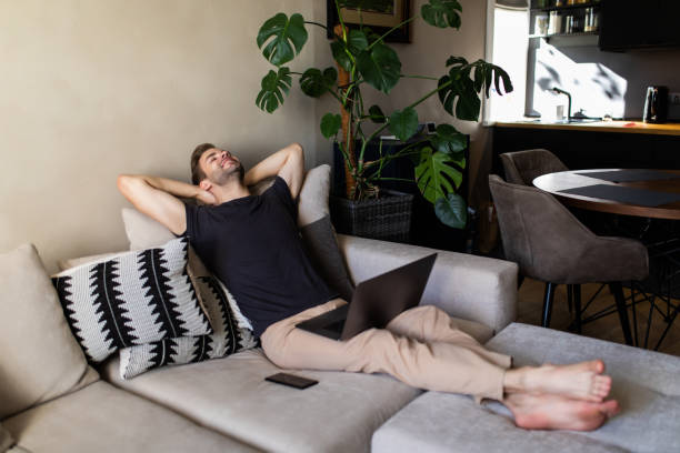 istock Smiling young man relaxing on a sofa with his laptop at home 1293487261