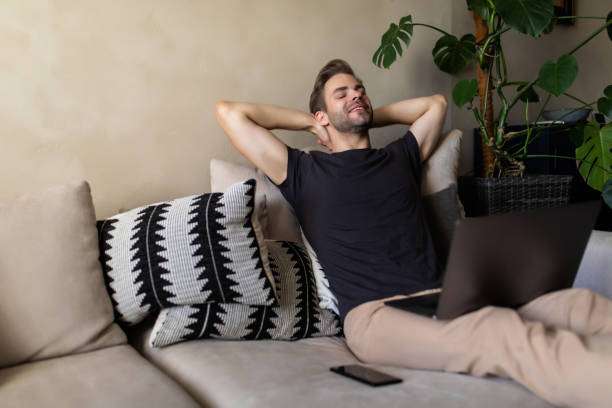 istock Smiling young man relaxing on a sofa with his laptop at home 1293487282