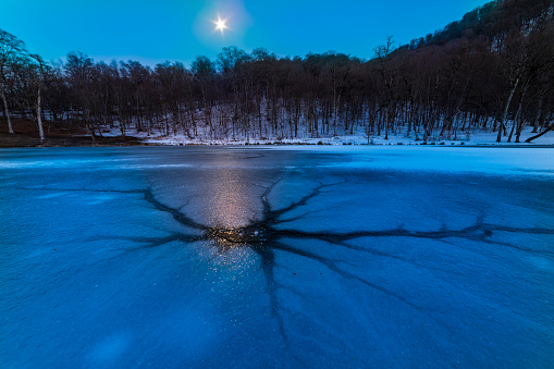 Beautiful winter night landscape. Frozen lake with crack on the ice, beautiful forest. Moon on the starry sky