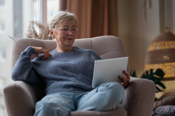 istock Senior woman with tablet pc resting on sofa near window reading something in internet. 1293500816