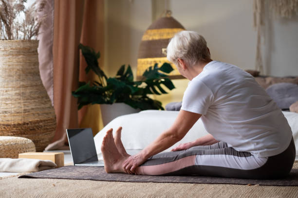 Sporty senior woman practicing yoga, doing seated forward bend pose, paschimothanasana Sporty senior woman practicing yoga, doing seated forward bend pose, paschimothanasana, Caterpillar in yin posture, working out at home. knee to the head pose stock pictures, royalty-free photos & images