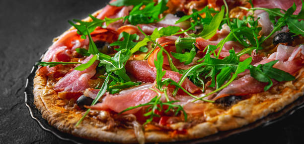istock Pizza with Mozzarella cheese, ham, pepper, olive, meat, Tomato sauce, Spices and Fresh arugula. Italian pizza on black plate on Dark grey black slate background 1293501067