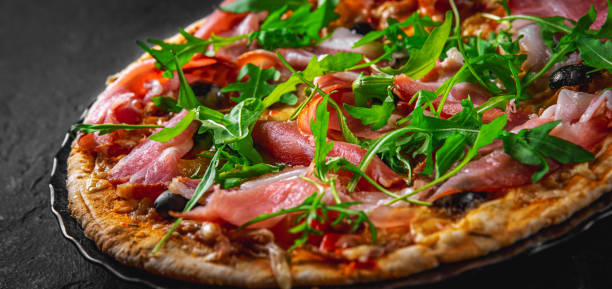 Pizza with Mozzarella cheese, ham, pepper, olive, meat, Tomato sauce, Spices and Fresh arugula. Italian pizza on black plate on Dark grey black slate background Pizza with Mozzarella cheese, ham, pepper, olive, meat, Tomato sauce, Spices and Fresh arugula. Italian pizza on black plate on Dark grey black slate background prosciutto stock pictures, royalty-free photos & images