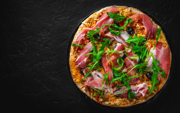 istock Pizza with Mozzarella cheese, ham, pepper, olive, meat, Tomato sauce, Spices and Fresh arugula. Italian pizza on black plate on Dark grey black slate background 1293501080