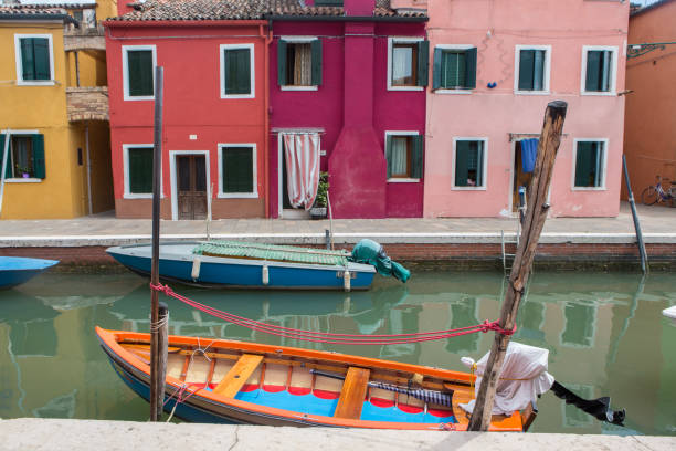 two small motor boats docked on the side of a canal that is surrouded by brightly painted houses on the island of burano which is near venice, italy. - rowboat nautical vessel small motorboat imagens e fotografias de stock