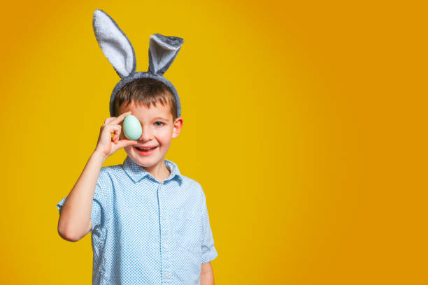 バニーの耳を持つ幸せな子供の男の子は黄色の背景にイースターエッグで彼の目を覆っています。黄色の背景にイースターエッグを持つ子供。 - easter egg easter yellow paint ストックフォトと画像