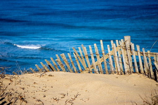 An old wooden fence protects a sand dune from erosion as a wave crashes in the background.