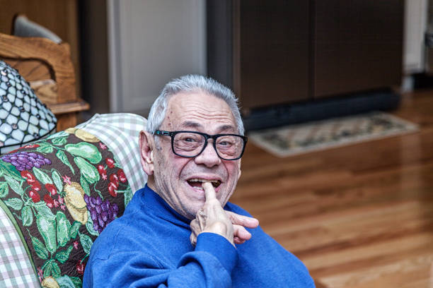 An elderly senior adult man is sitting at home in his living room looking at the camera. He is making an exaggerated funny squinting eyes face while biting the fingernail and end of his little pinky finger.