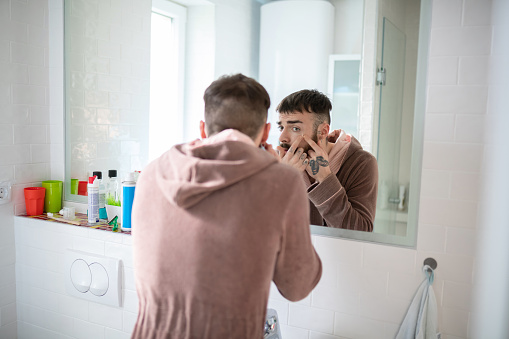 Young man in bathrobe in front of the bathroom mirror squeezing blackheads