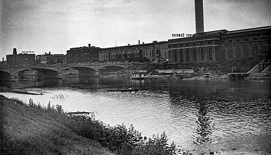 West bank of the Des Moines River from the Center Street Dam looking south in 1939. Features from right to left - water intake for electric plant, Des Moines Electric Company power plant with Des Moines Service sign, Grand Ave. Bridge, Des Moines Coliseum, a hotel, Des Moines Public library, Jack Sprat Foods cold storage warehouse for fruits and vegetables. Only the library building remains. Scanned film with soft focus and significant grain.