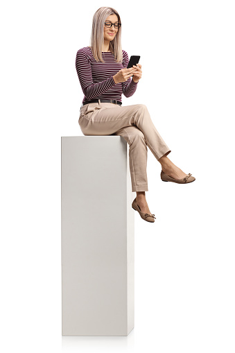 Professional young woman with glasses using a smartphone and sitting on a tall white column isolated on white background