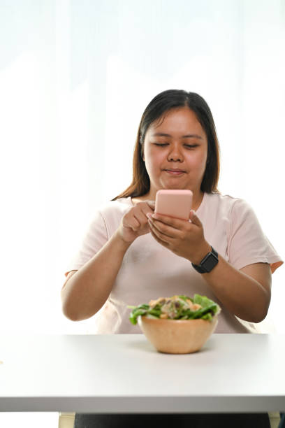 retrato de uma jovem obesa usando telefone inteligente tirando foto de salada em uma tigela em casa. - thai cuisine asian cuisine vertical close up - fotografias e filmes do acervo