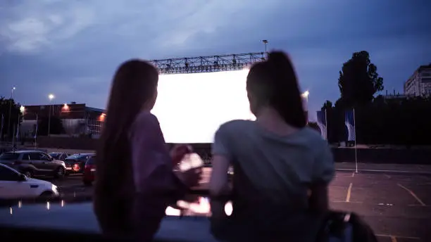 Rear view of two female friends sitting in the car while watching a movie in an open air cinema with a big white screen. Entertainment concept. Focus on a screen. Web Banner