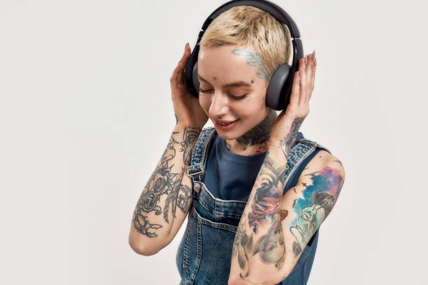 listening to music. a young white pierced and tattoed woman with her eyes closed and hands over her ears wearing a denim overall listening to music with her big earphones - headphones women tattoo music imagens e fotografias de stock