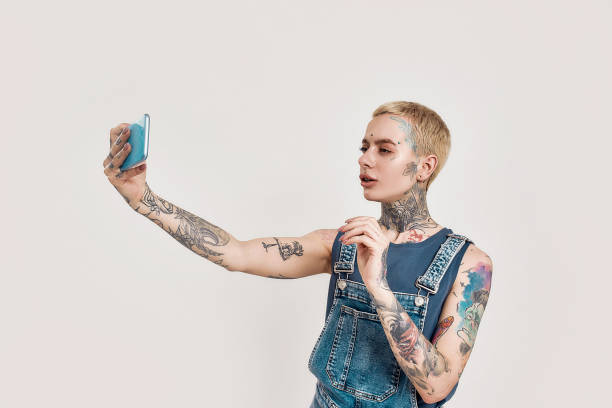 入れ墨とピアス。彼女のスマートフォンでかわいい自分撮りを作る全体的なデニムを身に着けている白い入れ墨のピアス女性 - tattoo t shirt brick wall portrait ストックフォトと画像