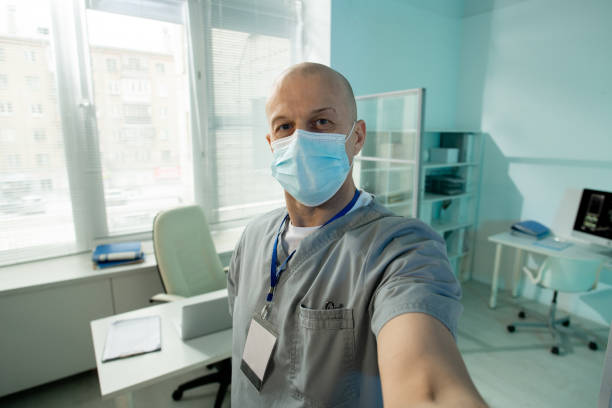 medico maschio maturo calvo in maschera protettiva che ti guarda mentre fai selfie - doctor male doctor mature men portrait foto e immagini stock