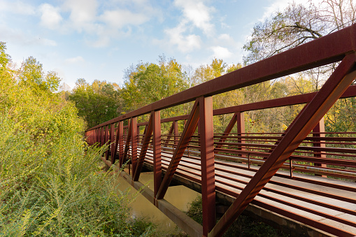 Side view of a red iron pedestrian bridge of a greenway surrounded by trees running over a river on the Neuse River Greenway in Raleigh, North Carolina, USA; landscape