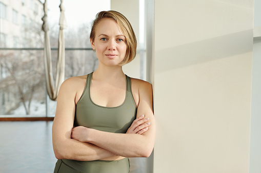 Happy young female fly yoga trainer in grey tracksuit keeping her arms crossed by chest while leaning by wall against hammock in gym