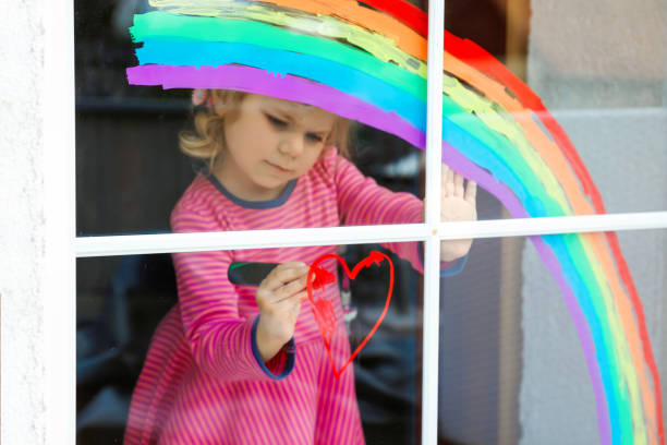 パンデミックコロナウイルス検疫中にカラフルな窓の色で塗装された虹を持つadoralbe小さな幼児の女の子。子供は世界中の虹を「みんな元気にしよう」という言葉で虹を描いています。 - epidemic paint virus illness ストックフォトと画像