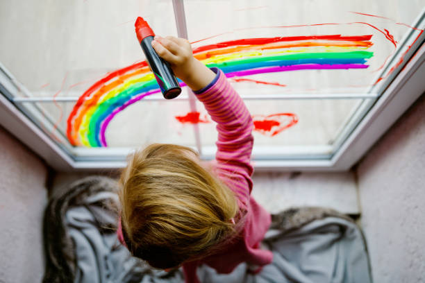 パンデミックコロナウイルス検疫中にカラフルな窓の色で描かれた虹と心を持つ愛らしい小さな幼児の女の子。世界中の子供の絵虹は、すべての井戸になろうという言葉で - epidemic paint virus illness ストックフォトと画像