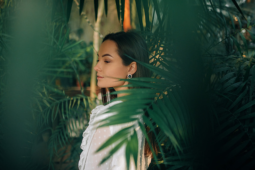 Portrait of young woman near big leaves of tropical plants inside orangery.