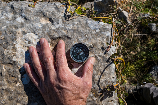Compass standing on rock. Hand surrounding the compass. Plants around.