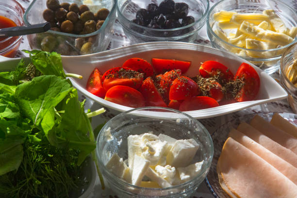 Homemade delicious, daily breakfast table at home. Thyme tomatoes, fresh arugula leaves, rosehip marmalade, cheese varieties, black and green olives, butter, smoked turkey slices, green and red sweet peppers. Horizontal close-up.