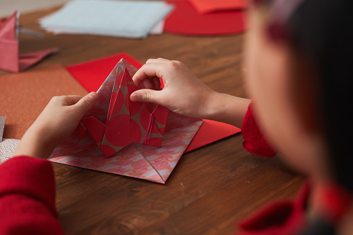 Unrecognizable little Asian girl sitting at wooden table making origami crane using colored paper, ocer-the-shoulder shot