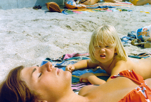 1976 vintage, seventies, retro colourful image of young girl looking up from her beach towel and a sunbathing woman in red bikini.