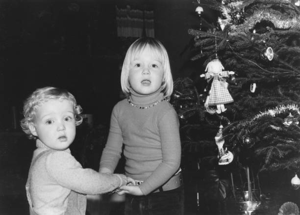Young siblings holding hands, standing next to a decorated christmas tree December 1977 vintage, retro monochrome image of a brother and sister, holding hands, standing next to a vintage decorated christmas tree. sibling photos stock pictures, royalty-free photos & images