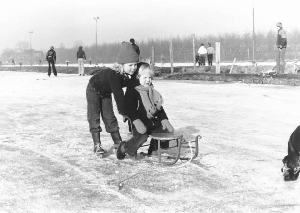 Vintage young girl in duffle coat on Frisian walkers pushing a boy on a retro sled on a natural ice rink. 1978 vintage, seventies, retro monochrome portrait of a young girl in duffle coat on Frisian walkers pushing a boy on a retro sled on a natural ice rink. ice skating photos stock pictures, royalty-free photos & images