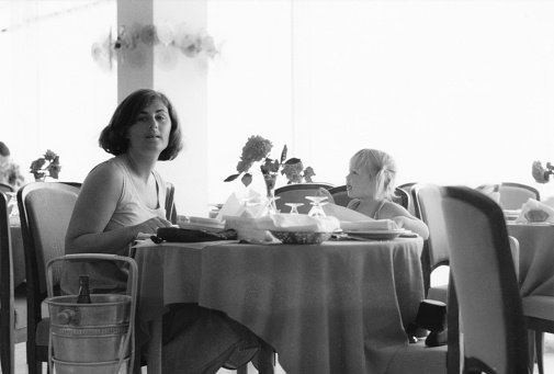 1976 vintage, seventies, retro monochrome image of a young mother and girl with blond hair sitting at a dining table in a restaurant of a hotel in Romania.